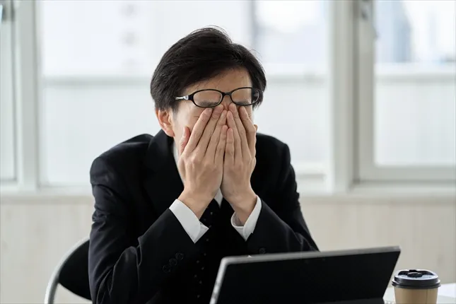 疲れた様子でパソコンの前に座り両手で顔を覆うスーツ姿の男性